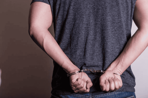 A person wearing a gray t-shirt and jeans with their hands handcuffed behind their back.